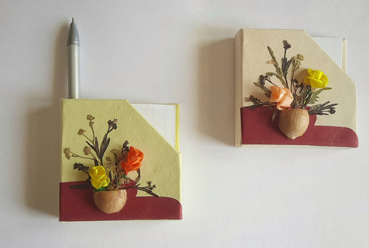 A magnetic chit holder with a floral design, featuring dry pressed leaves and seeds, displayed on a metallic surface.