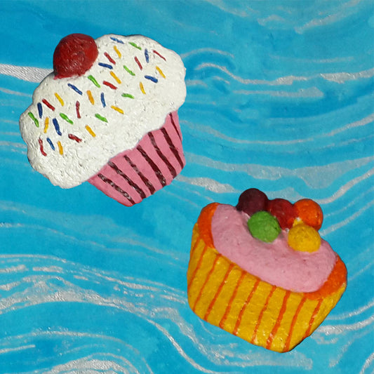 Hand-crafted paper pulp fridge magnets designed to look like cupcakes with colorful icing and sprinkles, and another resembling an ice cream with fruits on top, set against a blue background.
