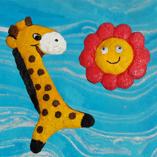 A hand-crafted paper pulp fridge magnet in the shape of a giraffe next to a flower-shaped magnet, both with vibrant colors and smiling faces.