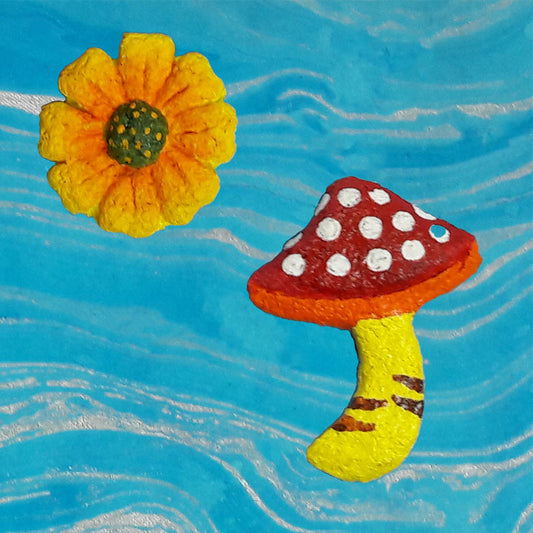 Handmade paper pulp fridge magnets designed to look like a yellow flower and a red and white spotted mushroom, placed against a blue background.