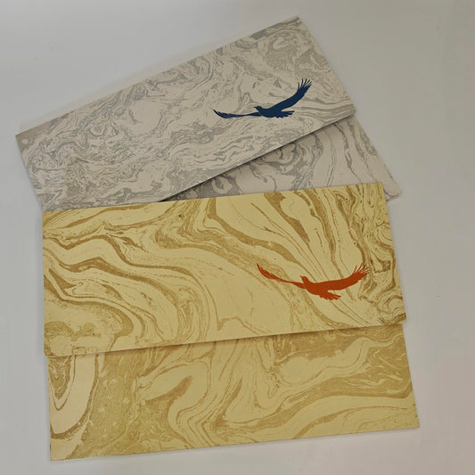Two decorative envelopes with marbled paper designs, one in beige and gray with a blue bird motif, and one in gold and beige with an orange bird motif, arranged on a light background.