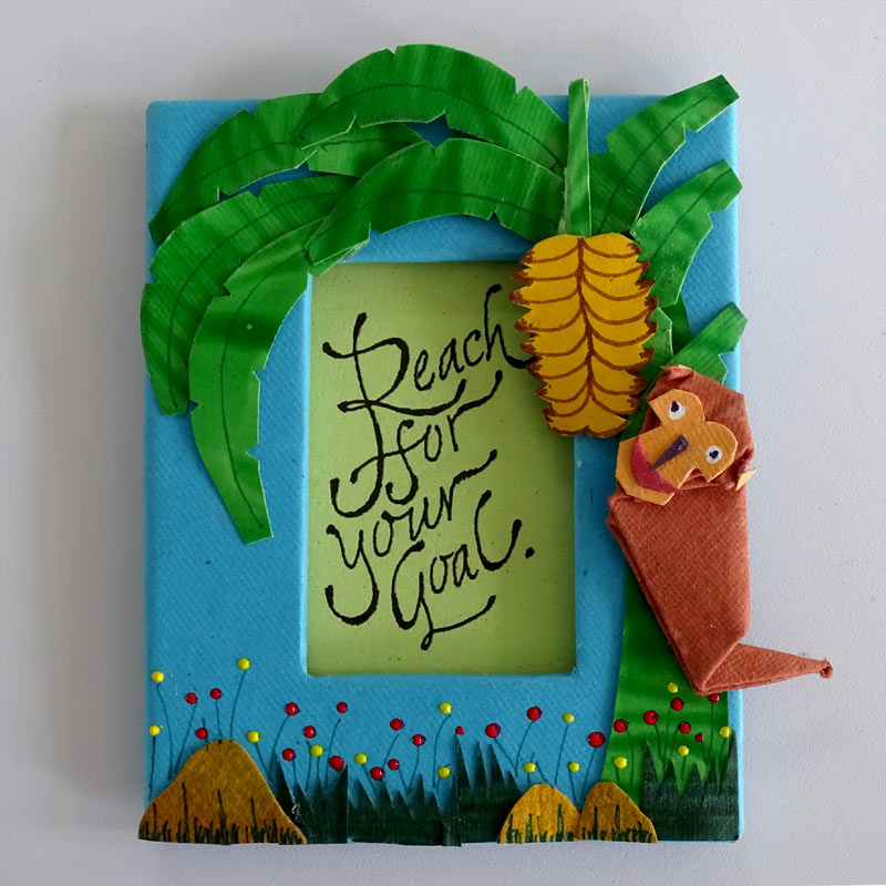 A Fridge Magnet with an origami Monkey on a paper cut-out banana tree & the motivational quote inscribed in a window in the center of the frame.
