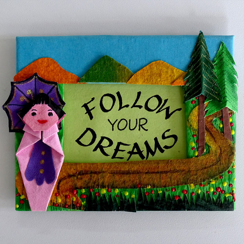 A Fridge Magnet with an origami Doll  with paper cut-out mountains, trees and a road. The motivational quote inscribed in a window in the center of the frame.