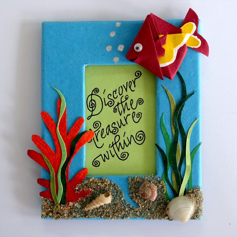 A Fridge Magnet with an origami Fish and paper cut-out corals  in deep sea with sand & shells & the quote inscribed in a window in the center of the frame.