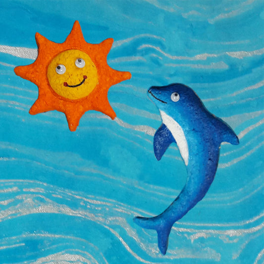 A hand-crafted paper pulp fridge magnet in the shape of a smiling sun with human-like features, placed above a magnet shaped like a dolphin in a blue ocean background.